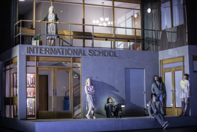 Näyttämölle rakennettu lavaste esittää koulurakennuksen sisäänkäyntiä ja sisätiloja, seinässä teksti "International School", ulko-oven liepeillä nojailee joukko nuoria, ylemmässä kerroksessa kaiteeseen nojaa keski-ikäinen opettajan näköinen nainen, kuva oopperasta Innocence.