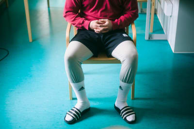 En fånge sitter på en stol. Axlar och huvud är inte med på bilden, men mannen är klädd i röd tröja, mörka shorts över gråa gymbyxor med strumporna uppdragna över byxorna. Han har händerna knäppta i famnen.