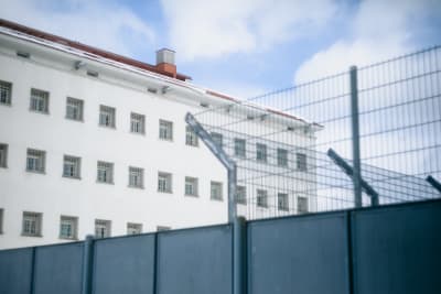 En ljus fängelsebyggnad med ett staket i förgrunden och rader av gallerförsedda fönster på andra sidan staketet.
