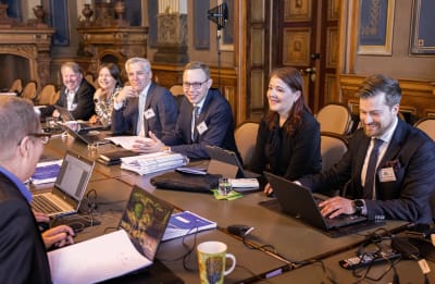En grupp politiker sitter vid ett avlångt bord inne i Statsrådet. De har bärbara datorer framför sig och pappershögar och är mitt i en diskussion.