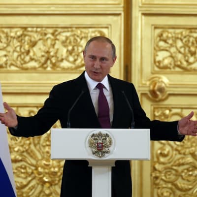 Putin puhuu korokkeen takaa ja levittää juuri käsiään. Putinilla on tumma asu ja viininpunainen kravatti. Taustalla näkyy suuri kullattu ja koristeltu ovi ja kuvan oikeassa ja vasemmassa reunassa Venäjän liput. Korokkeessa on Venäjän vaakunaeläin, kaksipäinen kotka.