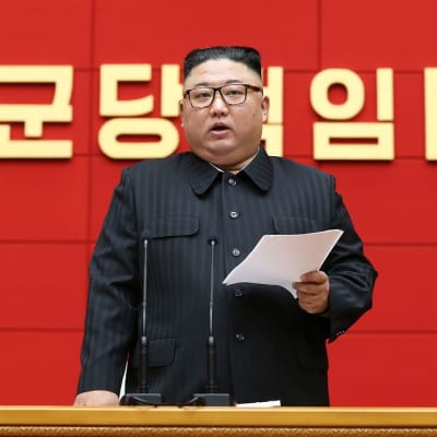 Kuvassa Kim Jong-un seisaallaan paperi kädessään. Taustalla seinässä on koreankielistä kirjoitusta. 