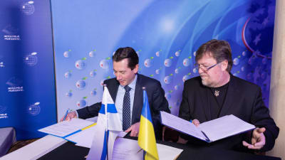 Jan D. Oker-Blom och Oleksandr Menzul sitter vid ett bord och undertecknar ett avtal. På bordet står Finlands och Ukrainas flaggor.