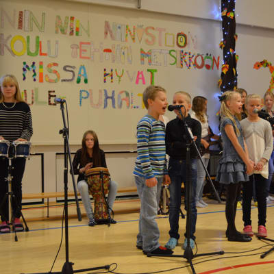 Oppilaat esiintyvät - laulavat ja soittavat koulun salissa.