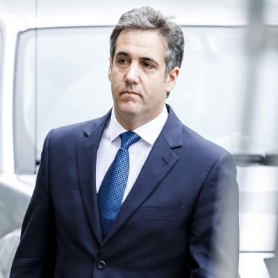 Cohen kävelee tummansinisessä puvussa, sininen kravatti kaulassaan, vakava ilme kasvoillaan.