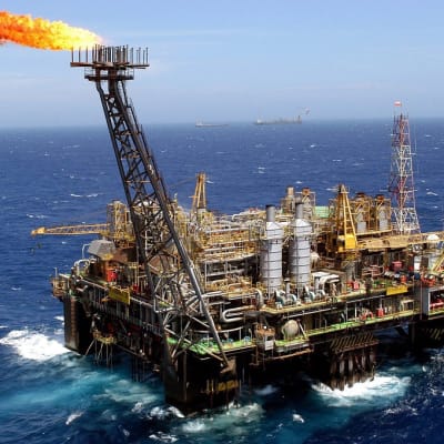 Brasilian Petrobras-yhtiön öljylautta merellä päivällä. Meri aaltoilee tummansinisenä, taivas on vaalea. Öljynporauslautan tornista nousee liekki, kun porauksen yhteydessä nousevaa kaasua poltetaan. Kaukana horisontissa näkyy kaksi alusta, mahdollisesti tankkereita.