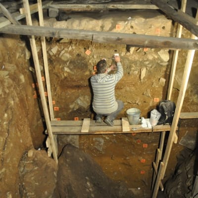 Telineellä kyyköttäjä tutkija kaivaa näytettä luolan pohjaan kaivetun kuopan seinämästä. 