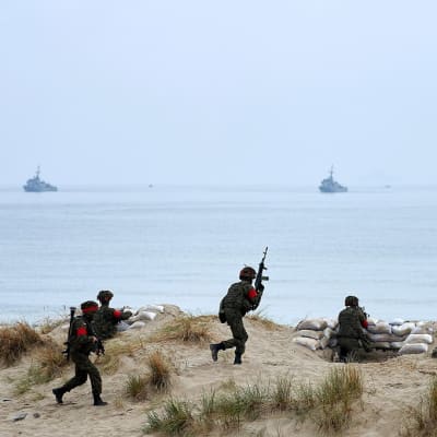 Sotilaat juoksevat hiekassa, taustalla meri jossa kaksi alusta.