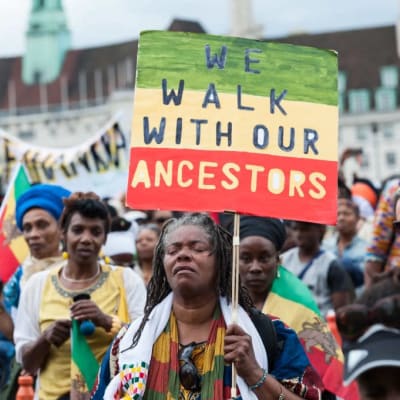 En kvinna går längst fram i en marsch med en skylt där det på engelska står "We walk with our ancestors"