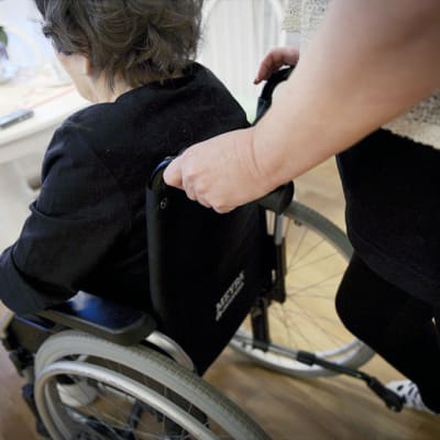 Omaishoitaja työntää vanhusta pyörätuolissa. 