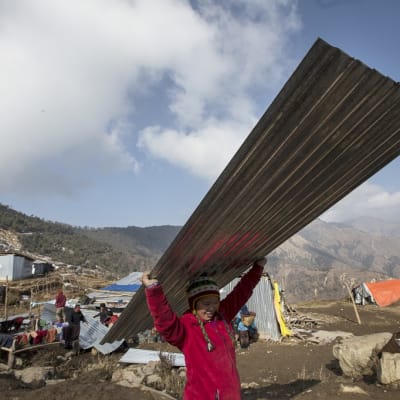 Nepalilainen tyttö kantaa aaltopeltiä päänsä yläpuolella. Tytöllä on kirkkaanpunainen takki ja pipo. Taustalla näkyy vuoristoista maisemaa ja väliaikaiselta näyttäviä rakennelmia. Kauempana pieni joukko ihmisiä on aaltopeltikasan luona.