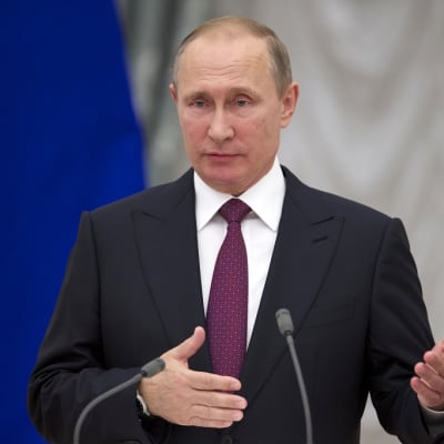 Putin puhuu edessään oleviin kahteen mikrofoniin. Hänellä on tumma puku, viininpunainen pilkullinen kravatti. Taustalla näkyy Venäjän lippua.