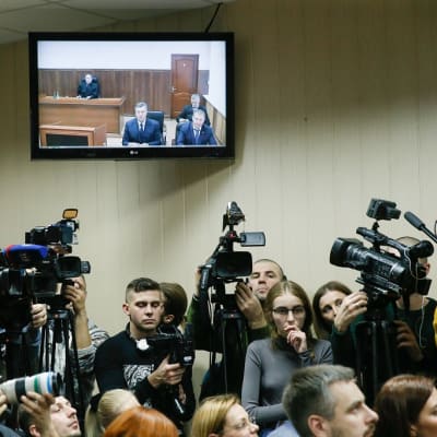 Toimittajien joukko seisoo televisioruudun alla. Ruudulla näkyy Ukrainan entinen presidentti rostovilaisessa tuomioistuimessa.
