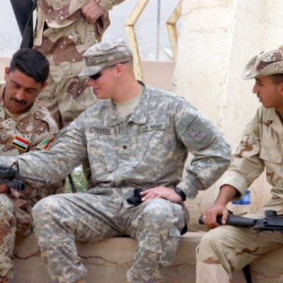Irakilaisten sotilaiden välissä istuu amerikkalaissotilas, asetta tutkitaan