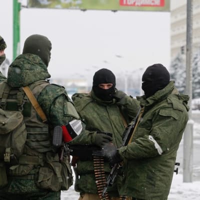 Neljä maastopukuista miestä kasvot peitettyinä ja aseet käsissään seisoo talvisella kadulla.