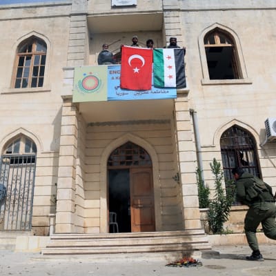 Mies juoksee kohti kivistä hallintorakennusta, jonka parvekkeelta miesjoukko roikuttaa Syyrian ja Turkin lippuja.
