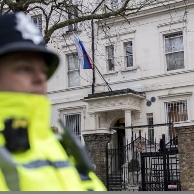Brittiläinen poliisimies seisoo keltaisissa huomioliiveissä, perinteinen bobby-hattu päässään vartiossa Venäjän vaalean suurlähetystörakennuksen edessä. Puissa ei ole lehtiä. Suurlähetystön julkisivulla on Venäjän lippu.