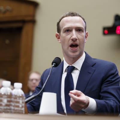 Tummansiniseen pukuun, valkoiseen kauluspaitaan ja tummansiniseen kravattiin pukeutunut Zuckerberg istuu pöydän takana ja puhuu mikrofoniin. Taustalla näkyy iso ovi. Pöydällä on kaksi vesipulloa.