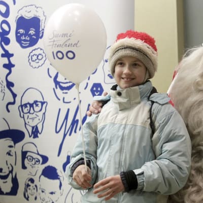 Suomi 100 -ilmapalloa pitelevä tonttulakkipäinen lapsi joulupukin kainalossa.