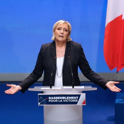 Marine Le Pen puhuu puhujanpöntön takana levittäen käsiään sivulle. Taustalla näkyy Ranskan lippuja.