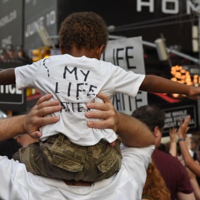 Pieni musta lapsi selin isänsä harteilla, paidan selässä lukee "my life matters".  