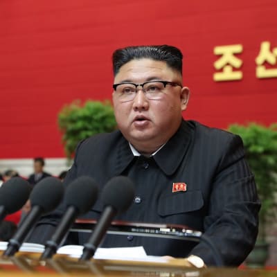 Kim Jong-un istuu pöydän takana. Hänen edessään on useita mikrofoneja. 