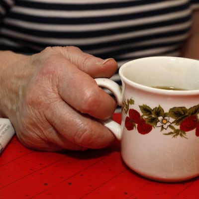 Ikääntyneen naisen käsi pitelee mansikkakuvioista kahvikuppia. Pöydällä näkyy myös sanomalehden kulma. 