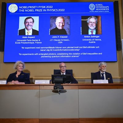 Voittajien kuvat valkokankaalla, edessä kolme Nobel-komitean edustajaa pöydän takana 