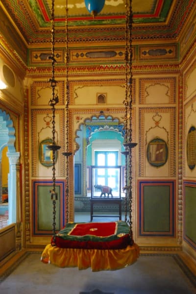 Indisk säng som hänger i taket med kättingar. Sängen utgörs av en röd-grön kvadratisk madrass av sammet.