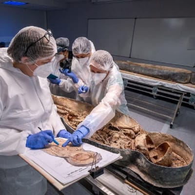 Neljä suojapukuista tutkijaa kumartuneena laboratoriossa lyijyarkussa olevan vainajan ylle. Tapana toinen arkku. 