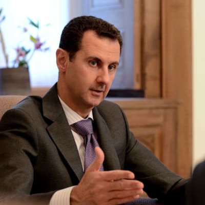 Syyrian presidentti Bashar al-Assad puhumassa ranskalaisille toimittajille. Assad selittää jotain tehostaen sanoja nostamalla vasenta kättään. Viiksekkäällä Assadilla on tummanharmaa puku, violetti ruutukuvioinen kravatti ja vaalea kauluspaita, jossa on raitoja. Taustalla on ikkunalauta, jolla on kukkaruukku.