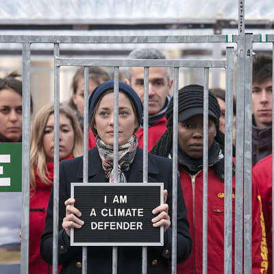 Ranskalainen näyttelijätär Marion Cotillard seisoo häkissä vierellään punatakkisia ihmisiä. Kädessään hänellä on kyltti, jossa lukee: "I AM A CLIMATE DEFENDER".