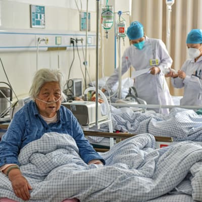 Kaksi koronapotilasta kiinalaisessa sairaalassa. Edessä istuu vuoteessaan vanha nainen, jolla on happiviikset. Takana on kaksi suojavarusteisiin pukeutunutta hoitajaa toisen potilaan vuoteen ääressä. 
