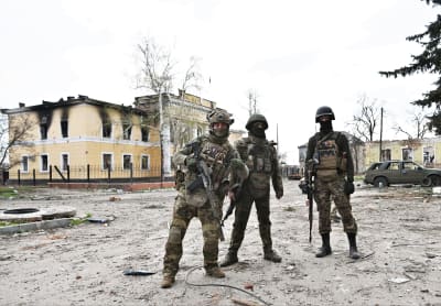 Tre soldater tittar in mot kameran, en av dem håller upp tummen. De står vid ett förstört hus.