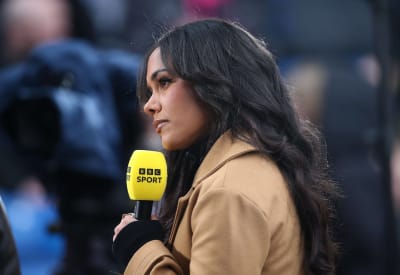 Alex Scott is photographed from the side.  She looks forward and holds a yellow microphone with text "BBC Sports".  She is wearing a beige coat.