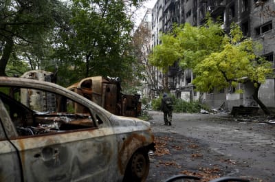 En bil som brunnit upp står bredvid ett skadat bostadshus medan kriget mellan Ryssland och Ukraina fortsätter. Andra förstörda bilar syns, en man går bort från bilden. Ett grönt träd växer framför flervåningshuset.