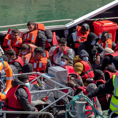 Kuvassa miehiä istumassa laivan kannella pelastusliivit päällään. 