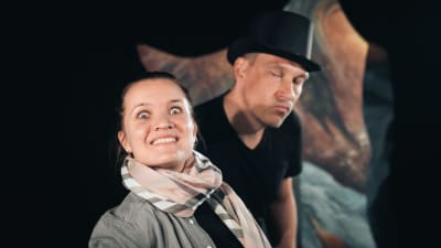 En kvinna och en man står på en teaterscen och ser in i kameran. Mannen har en hög hatt på huvudet.