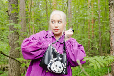 YleX:n Katri Norrlin tutkii omaa käsilaukkuaan metsässä. Norrlin katsoo vasemmalle salaperäisen näköisenä.