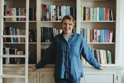 Katarina Sehm-Patomäki iklädd en jeansskjorta poserar framför en vit bokhylla fylld med böcker.