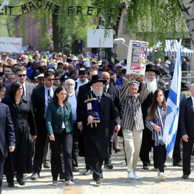 Ihmisjoukko alittaa portin, jossa lukee "Arbeit macht frei". Kaksi kantaa Israelin lippua, rabbi juutalaista kirjakääröä.