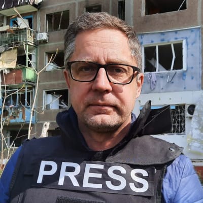 Redaktör Antti Kuronen framför ett sönderskjutet höghus. Antti är klädd i en skyddsväst med texten "Press".