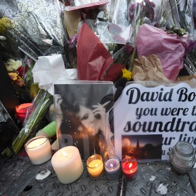 Keko muovi- ja paperikääröissä olevia kukkia ja niiden edessä kyntilöitä. Bowien valokuvan vieressä iso kortti, jossa lukee englanniksi: "David Bowie, olit elämämme elokuvan ääniraita".