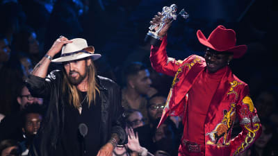 En svart man i en röd cowboyutstyrsel. I handen har han en vinststatyett. Brevid honom står en vit man i cowboyhatt och rockigt utseende.