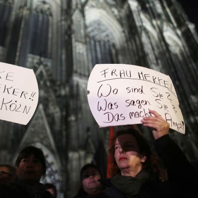 Kaksi naista pitelee saksankielisiä kylttejä. Toisessa lukee: "Kiitos, rouva Reker. Köln-parka." Toisen teksti kuuluu: "Rouva Merkel. Missä olette? Mitä sanotte? Tämä pelottaa." Naisten taustalla on Kölnin katedraali.