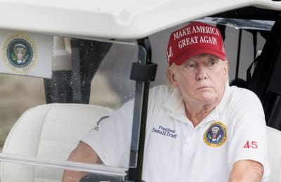 Donald Trump i en golfbil iklädd en vit t-tröja med krage och siffran 45 på ärmen. På ena bröstet står det President Donald Trump och på det andra finns en bild av USA:s presidentsigill. På huvudet har Trump en röd Make America Great Again-keps.