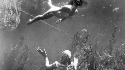 Suomuinen humanoidi hirviö katselee järven pohjakasvillisuuden lomasta yllä uivaa uimaasuista naista.