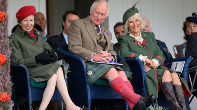 Prinsessan Anne, prins Charles och Camilla, hertiginnan av Cornwall, sitter i blå korgstolar och ser nöjda ut. Damerna är klädda i kappor i grönt, Anne har en röd hatt som matchar knapparna på hennes kappa och och Camilla har en grön hatt som matchar kappan. Charles är klädd i kavaj och kilt med röda knästrumpor.