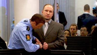 Polis öppnar Breiviks handbojor i rättegångssalen, åskådare tittar på.