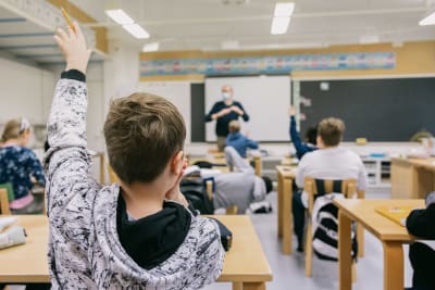 En elev markerar i ett klassrum. I bakgrunden syns lärare framför tavlan. 